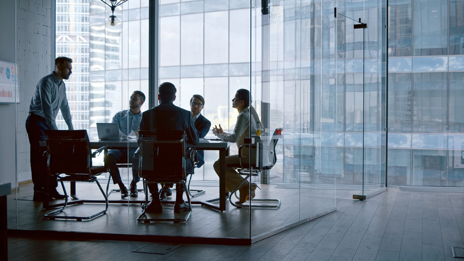 Business meeting in a modern glass-walled conference room with five people discussing.